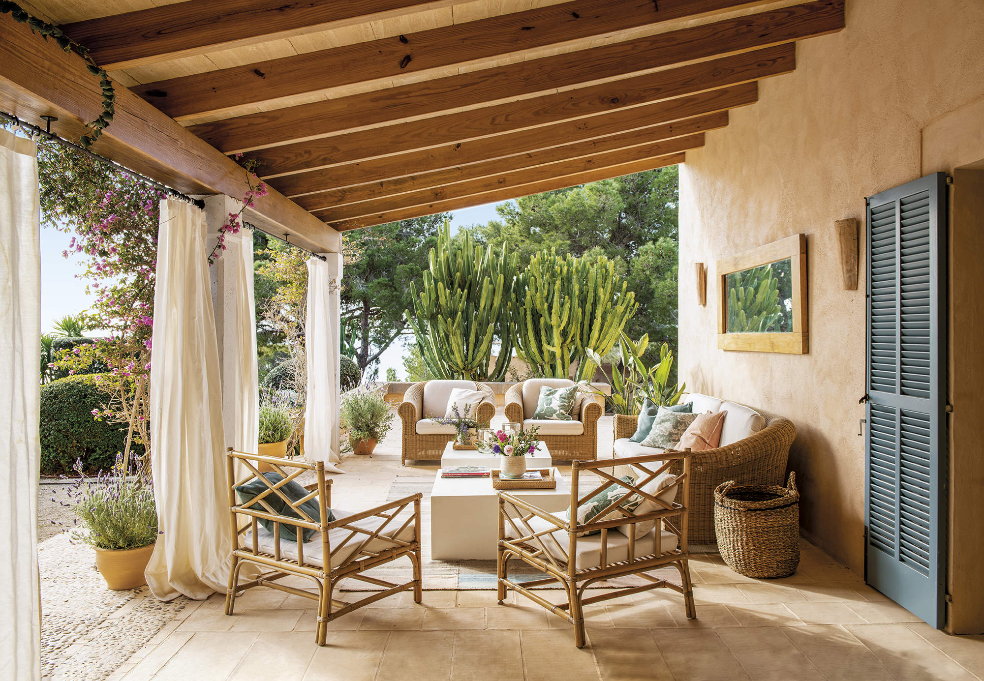 porche con suelo de barro, sofás de fibra con colchoneta blanca, butacas de cañan y dos mesas de centro blancas, techo con vigas y cortina blanca, plantas, cactus, lavanda